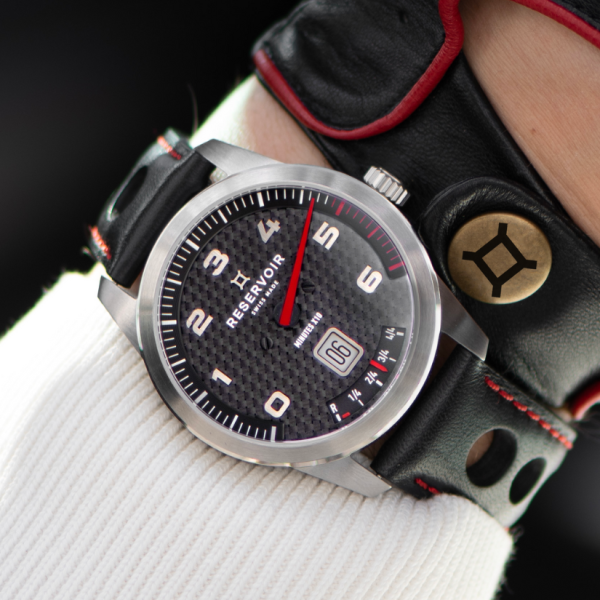 Wrist wearing a stainless steel watch with black dial, white numerals, red minute hand, and date display.