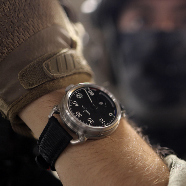 Tactical-gloved person wearing a wristwatch with a black dial and nylon strap, blurred background.