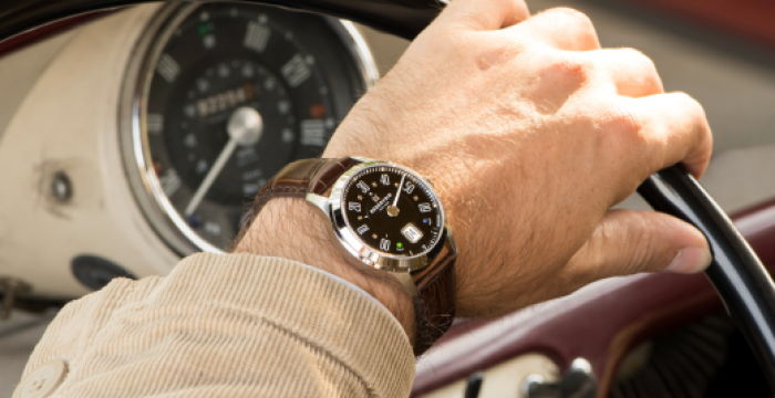 A left hand on a steering wheel wearing a wristwatch in a vintage car with visible dashboard gauges.