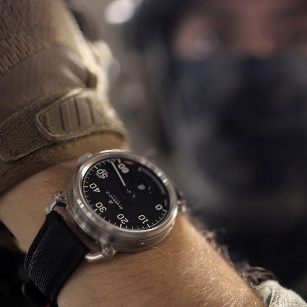 Wristwatch with black dial and strap on wrist, blurred background with person in military gear.