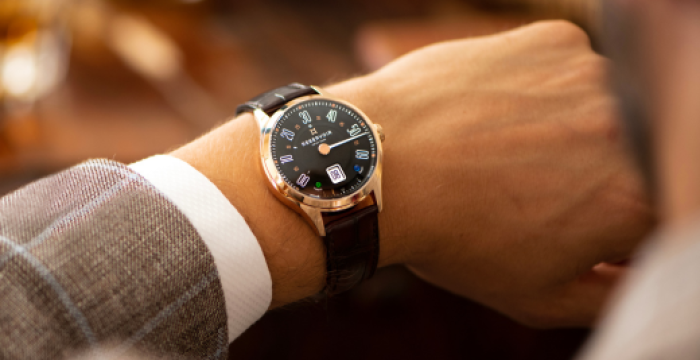 Person wearing a wristwatch with black dial, blue numerals, date window, and brown leather strap.